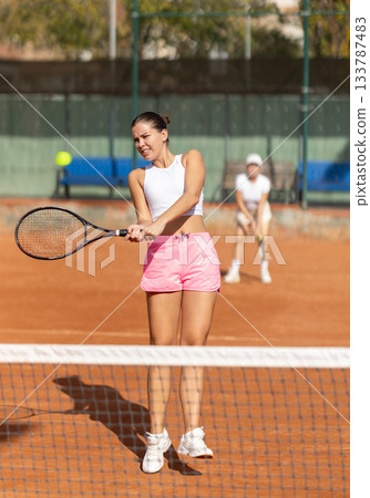 Two women in sportswear spending time playing sports on tennis courts, young girl receiving hit from tennis racket Two women in sportswear spending time playing sports on tennis courts, young girl receiving hit from tennis racket 133787483