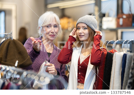 Young woman is fitting on and trying on warm hat in store, consulting with senior female assistant. Young woman is fitting on and trying on warm hat in store, consulting with senior female assistant. 133787546