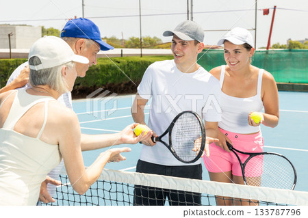 Group of tennis players standing on open court on warm day with rackets and balls in hands, friendly chatting after match 133787796