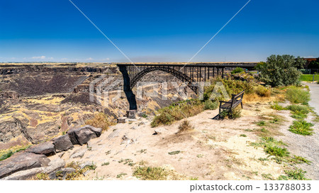 A view of the Perrine Bridge arch in Twin Falls, Idaho. The steel structure spans the deep basalt cliffs of the Snake River Canyon under a clear blue sky. Represents engineering and infrastructure 133788033