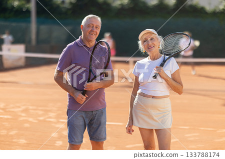 Elderly couple man and woman in sportswear posing with tennis rackets on tennis court after match Elderly couple man and woman in sportswear posing with tennis rackets on tennis court after match 133788174