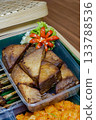 Close-up photo of tahu bacem (Indonesian braised and fried tofu) in a plastic tray, featuring caramelized triangles and a carved chili garnish on banana leaf 133788536