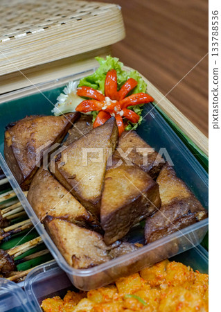 Close-up photo of tahu bacem (Indonesian braised and fried tofu) in a plastic tray, featuring caramelized triangles and a carved chili garnish on banana leaf 133788536