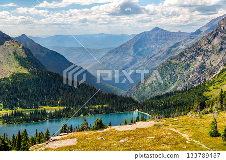 Hidden Lake sits in deep valley in Glacier National Park Montana. Alpine landscape features blue water and green pine forests surrounded by rugged mountain peaks 133789487