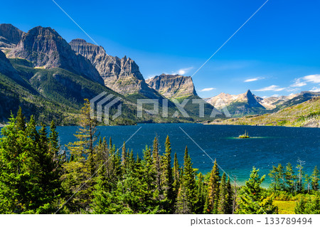 Panoramic view of the vibrant blue Saint Mary Lake, featuring Wild Goose Island, in Glacier National Park. The Rocky Mountains rise grandly under a clear blue sky, framed by pine trees Panoramic view of the vibrant blue Saint Mary Lake, featuring Wild Goose Island, in Glacier National Park. The Rocky Mountains rise grandly under a clear blue sky, framed by pine trees 133789494