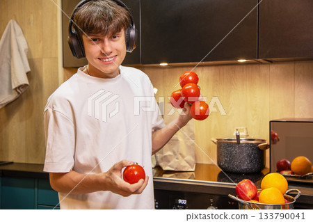 Cheerful teenager wearing headphones holding red tomatoes for making healthy salad. Concept of healthy lifestyle, cooking at home and listening to music while doing housework. Cheerful teenager wearing headphones holding red tomatoes for making healthy salad. Concept of healthy lifestyle, cooking at home and listening to music while doing housework. 133790041