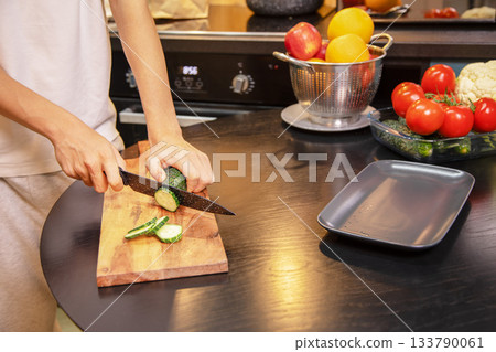 Person cutting fresh cucumbers on a wooden board surrounded by vegetables and fruits on the kitchen table. Healthy salad preparation and clean eating lifestyle at home. Person cutting fresh cucumbers on a wooden board surrounded by vegetables and fruits on the kitchen table. Healthy salad preparation and clean eating lifestyle at home. 133790061