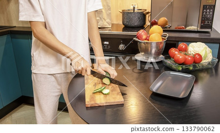 Person cutting fresh cucumbers on a wooden board surrounded by vegetables and fruits on the kitchen table. Healthy salad preparation and clean eating lifestyle at home. Person cutting fresh cucumbers on a wooden board surrounded by vegetables and fruits on the kitchen table. Healthy salad preparation and clean eating lifestyle at home. 133790062