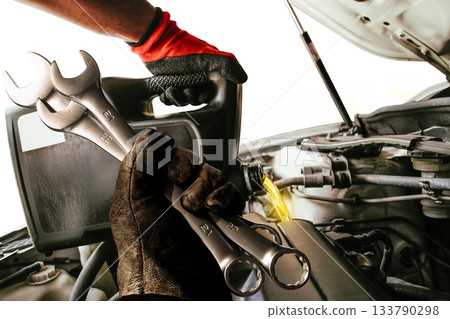 Mechanic in gloves pouring oil into a car engine while holding wrenches 133790298