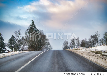 Snowy Road Scene With Wet Asphalt, Winter Trees, And Open Horizon Under Cloudy Sky Snowy Road Scene With Wet Asphalt, Winter Trees, And Open Horizon Under Cloudy Sky 133790312