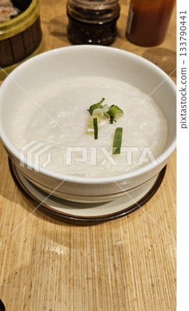 A warm bowl of congee garnished with green onions sits on a wooden table, surrounded by savory condiments, offering a comforting and customizable taste of traditional Asian cuisine. 133790441