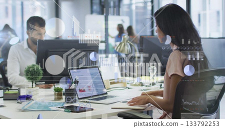 Typing woman in blush blouse analyzing report on laptop in office, with notebook, monitors, plant 133792253