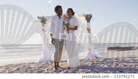 Standing bride and groom in wedding attire holding bouquets on sandy beach with floral arch 133792271