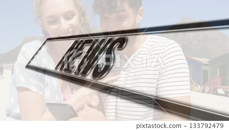 Smiling couple sharing smartphone on sand, with colorful wooden beach huts and NEWS graphic overlay 133792479