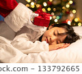 A child sleeps, eagerly waiting for a Christmas present. Santa Claus quietly places a present next to the child's pillow. 133792665