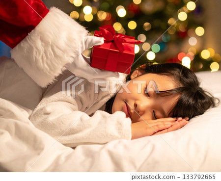 A child sleeps, eagerly waiting for a Christmas present. Santa Claus quietly places a present next to the child's pillow. 133792665
