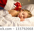 A child sleeps, eagerly waiting for a Christmas present. Santa Claus quietly places a present next to the child's pillow. 133792668