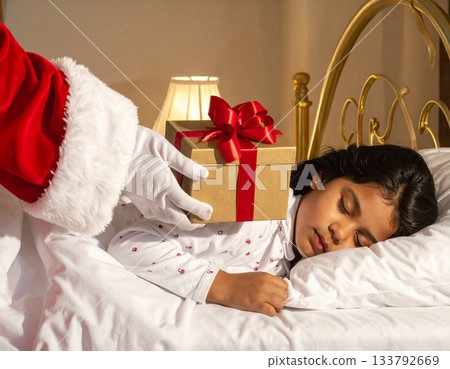 A child sleeps, eagerly waiting for a Christmas present. Santa Claus quietly places a present next to the child's pillow. 133792669