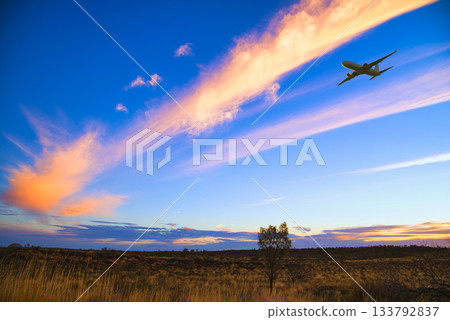 夕陽下的雲彩橫跨荒野，一架飛機穿梭其中。 133792837