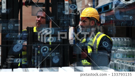 Two men wearing vests, helmet operating forklift controls in warehouse with shelving and pallets 133793135
