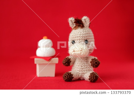 一個鉤針編織的小馬玩偶坐在一個鏡餅旁邊。 一個鉤針編織的小馬玩偶坐在一個鏡餅旁邊。 133793370