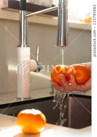 Person Washes Two Mandarin Oranges Under Running Water in Kitchen Sink 133795662