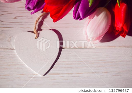 Flat design white wooden heart ornament is resting on tabletop with red, pink, purple tulip bouquet Flat design white wooden heart ornament is resting on tabletop with red, pink, purple tulip bouquet 133796487
