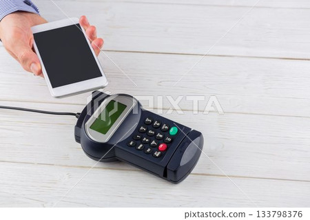 Man is holding smartphone above POS terminal with cable on pale wooden tabletop in flat design 133798376