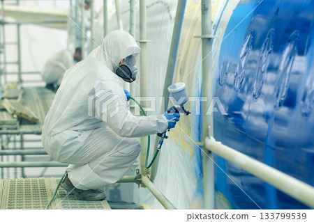 Worker paints aircraft surface in hangar during maintenance, demonstrating skills while ensuring quality finish and safety in a controlled environment Worker paints aircraft surface in hangar during maintenance, demonstrating skills while ensuring quality finish and safety in a controlled environment 133799539
