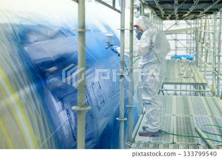Worker applies blue paint to large industrial tank using a spray gun while standing on scaffolding in a well-lit workspace during daylight hours Worker applies blue paint to large industrial tank using a spray gun while standing on scaffolding in a well-lit workspace during daylight hours 133799540