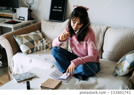 Young adult woman sits on sofa contemplating personal goals while looking at open notebook on coffee table. Goal setting, New Year resolutions, self reflection, mental health. 133801741