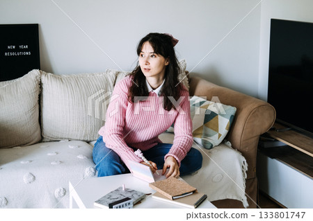 Young adult woman sits on sofa writing goals in small notebook next to letter board displaying NEW YEAR RESOLUTIONS. New Year resolutions, abundance mindset, wealth goals, money manifestation. 133801747