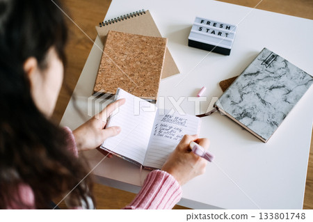 Person writes New Year resolutions list in small notebook on white table next to FRESH START light box. New Year resolutions, mindful goals, productivity tools, journaling for success. 133801748