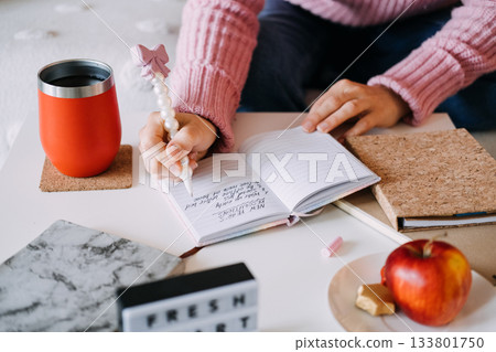 Person writes New Year resolutions list in small notebook using pearl pen next to coffee tumbler and apple. New Year resolutions, goal setting, journaling, healthy lifestyle. Person writes New Year resolutions list in small notebook using pearl pen next to coffee tumbler and apple. New Year resolutions, goal setting, journaling, healthy lifestyle. 133801750