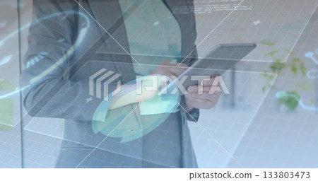 Holding tablet, executive in gray suit using holographic UI in office with grid charts plus icons Holding tablet, executive in gray suit using holographic UI in office with grid charts plus icons 133803473