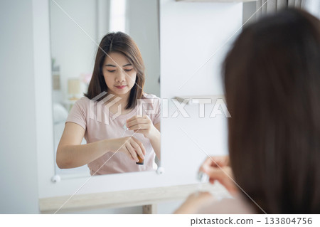 Young Woman Applying Skincare Product in Modern Bathroom Mirror Reflection, Emphasizing Self-Care and Daily Beauty Routine 133804756