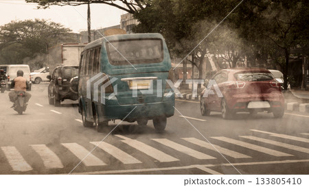 City street with vehicles emitting exhaust fumes. A blue van is in the foreground, with a red car and a motorcycle nearby. Concept of air pollution 133805410