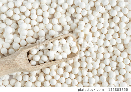 White dry tapioca pearls in wooden scoop on tapioca background. Top view of raw tapioca starch balls used for bubble tea or desserts. Tapioca pearls as healthy gluten free ingredient. 133805866