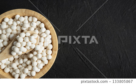 Organic tapioca pearls in wooden bamboo bowl on black table top view with copy space. Spoon scooping white dry tapioca pearls, cooking ingredient, gluten free food, healthy eating. 133805867