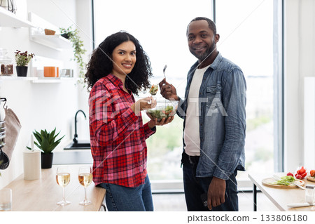 A smiling couple enjoys a healthy salad in a bright, modern kitchen setting. They are holding salad bowls and forks. 133806180