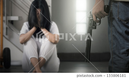 Human hand holding a revolver handgun in front of a woman in distress, sitting on the floor with her face hidden. Concept of gun violence 133806181