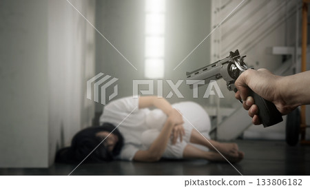 Human hand holding a revolver handgun in front of a woman in distress, sitting on the floor with her face hidden. Concept of gun violence 133806182