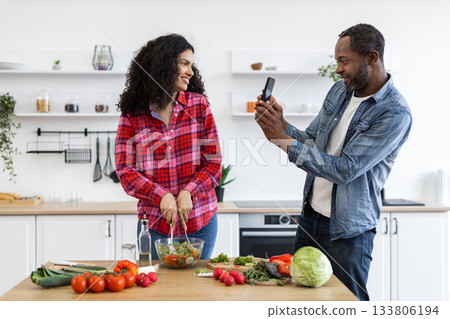 A couple is in the kitchen, with the woman preparing a salad while the man takes a photo with his phone. 133806194
