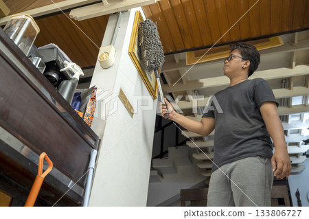 Indonesian southeast asian man is cleaning dust from a picture frame using a duster. Cleaning home activity Indonesian southeast asian man is cleaning dust from a picture frame using a duster. Cleaning home activity 133806727