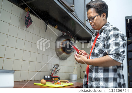 Indonesian southeast asian man standing in a kitchen peeling a red onion with a knife. Preparing ingredients on a cutting board 133806734
