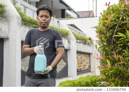 Indonesian southeast asian gardener man wearing white gloves watering a potted plant in the backyard. Gardening work Indonesian southeast asian gardener man wearing white gloves watering a potted plant in the backyard. Gardening work 133806740