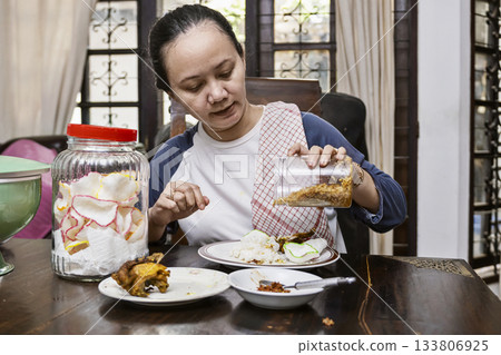 Indonesian woman enjoying having meal in dining table. She is putting fried onion on the plate to eat 133806925