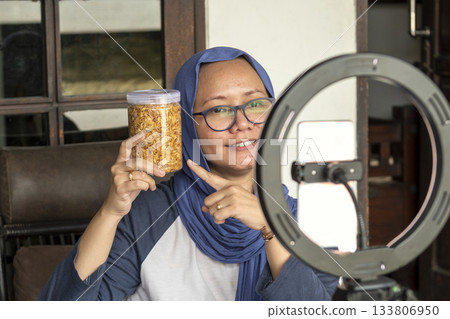 Indonesian hijab woman is pointing a jar of food and doing livestream for selling at home. Indonesian hijab woman is pointing a jar of food and doing livestream for selling at home. 133806950
