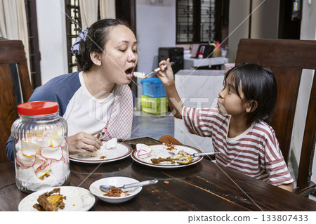 Asian woman and a child are eating in dinning table. Daughter feeding her mother while eating together. Asian woman and a child are eating in dinning table. Daughter feeding her mother while eating together. 133807433