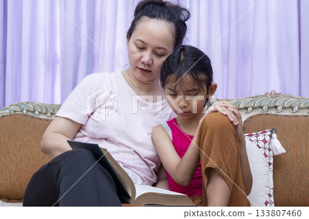 Woman and a child are sitting on a couch reading a book together 133807460